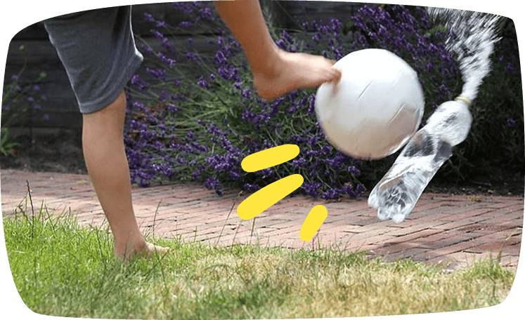 A person kicks a white ball barefoot on a brick patio, sending a plastic bottle flying nearby, with purple lavender bushes in the background.