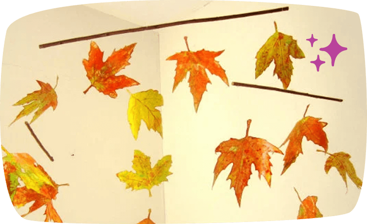 A decorative autumn mobile made from colorful fall leaves in shades of red, orange, and yellow, suspended from wooden sticks against a cream-colored background.