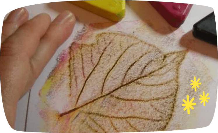 A child's hand holds down a leaf on paper while creating a leaf rubbing art project using colorful wax crayons or pastels.