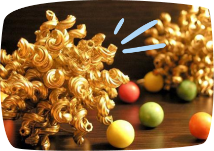 Two golden decorative sculptures made from curly pasta sit on a wooden surface, surrounded by small colorful balls in red, yellow, and green.