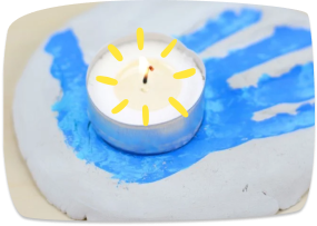 A child's blue handprint painted on a white surface with a small tealight candle decorated with yellow rays to look like a sun placed in the center.