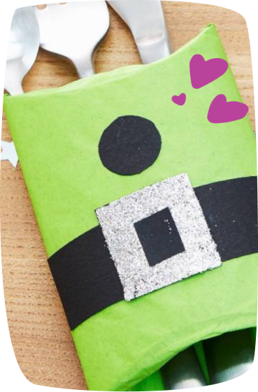 A green gift wrapped to look like a leprechaun, featuring a black belt with a glittery silver buckle and pink hearts, perfect for St. Patrick's Day.