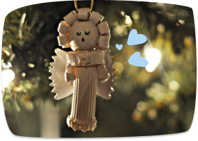 A handmade straw angel Christmas tree ornament hanging on a decorated tree, with small blue heart graphics overlaid on the image.