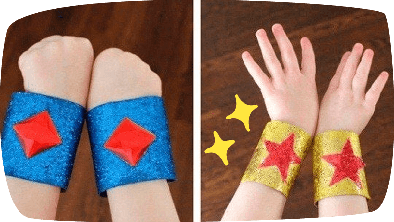 Two pairs of children's wrists wearing sparkly DIY superhero cuffs, one pair in blue glitter with red diamond gems and another in gold glitter with red stars.