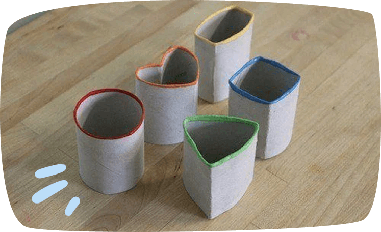 Five toilet paper rolls reshaped into different forms — a circle, heart, triangle, square, and rectangle — with colorful painted rims, displayed on a wooden surface.